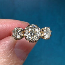 Load image into Gallery viewer, A three-stone diamond ring held between fingers against a dark blue background. The center stone is round brilliant-cut, flanked by two smaller round stones, set in a yellow gold band.