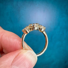 Load image into Gallery viewer, A close-up of a gold ring with three small clear stones held between fingers against a textured blue background.