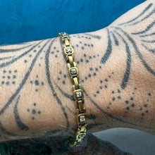 Load image into Gallery viewer, A gold bracelet with clear gemstones worn on a tattooed forearm, set against a textured blue background.
