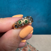 Load image into Gallery viewer, A close-up of a hand with peach-colored nail polish holding a vintage-style gold ring decorated with green and blue gemstones.
