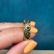 Load image into Gallery viewer, A gold ring with green and blue gemstones, held between fingers with orange nail polish against a textured blue background.
