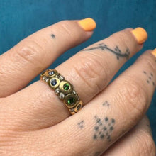 Load image into Gallery viewer, A gold ring with three green oval stones and small clear stones, worn on a tattooed hand with orange nail polish against a textured blue background.
