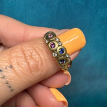 Load image into Gallery viewer, A gold band ring with small colorful gemstones, held on a person's finger against a textured teal background.
