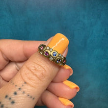 Load image into Gallery viewer, A gold ring with multiple small colorful gemstones, worn on a finger with orange nail polish, set against a textured teal background.

