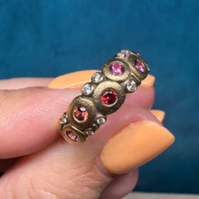 Load image into Gallery viewer, A hand with peach-colored nails holds a vintage-style gold ring decorated with round red and pink gemstones and small diamonds, set against a textured blue background.
