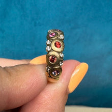 Load image into Gallery viewer, A gold ring with three round red gemstones and small clear stones, held between fingers against a textured blue background.
