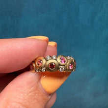 Load image into Gallery viewer, A hand with yellow-orange painted fingernails holds a gold band ring decorated with small diamonds and three round pink gemstones, set against a textured teal-blue background.
