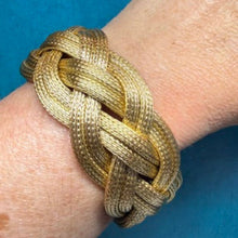Load image into Gallery viewer, A close-up of a person's wrist wearing a braided gold-toned metal cuff bracelet against a textured blue background.
