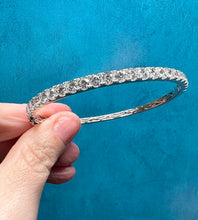 Load image into Gallery viewer, A silver-toned diamond tennis bracelet held by a hand against a textured blue background.
