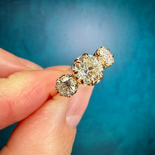 Load image into Gallery viewer, A close-up of a hand holding a small gold diamond ring with three round-cut diamonds set in a row, against a textured teal blue background.
