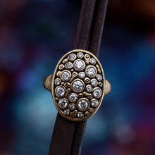 Load image into Gallery viewer, A close-up of a vintage-style oval ring with a gold-toned frame, set with multiple clear round stones, worn on a dark brown leather strap against a blurred multicolor background.
